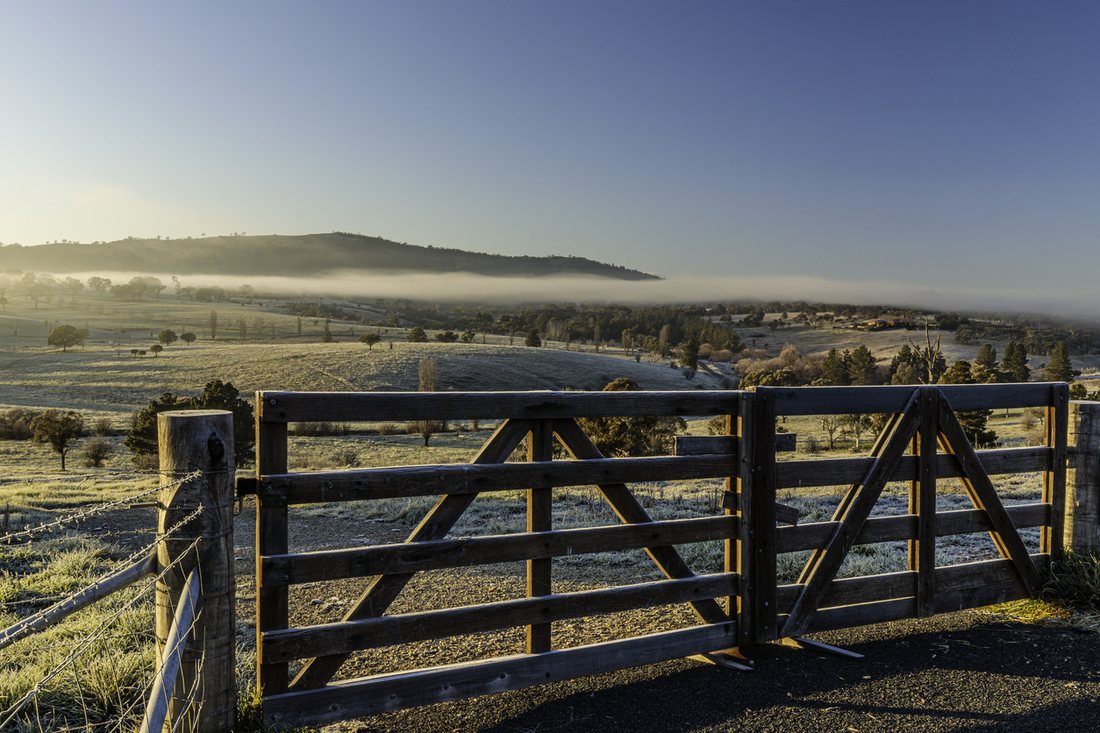 A closed timber farm gate at dawn, with frost on the paddocks and fog in the valley beyond